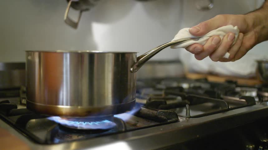 A mans hand moving a saucepan over a stove gas burner as steam rises from the contents