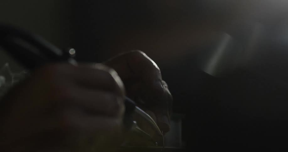 In a darkened work space a person can be seen carefully using a soldering gun as smoke wafts up from the soldering