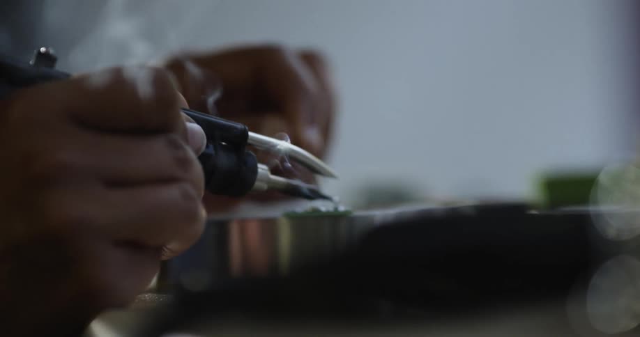 A man carefully holds a soldering gun and works on a metal object while smoke wafts up from the soldering