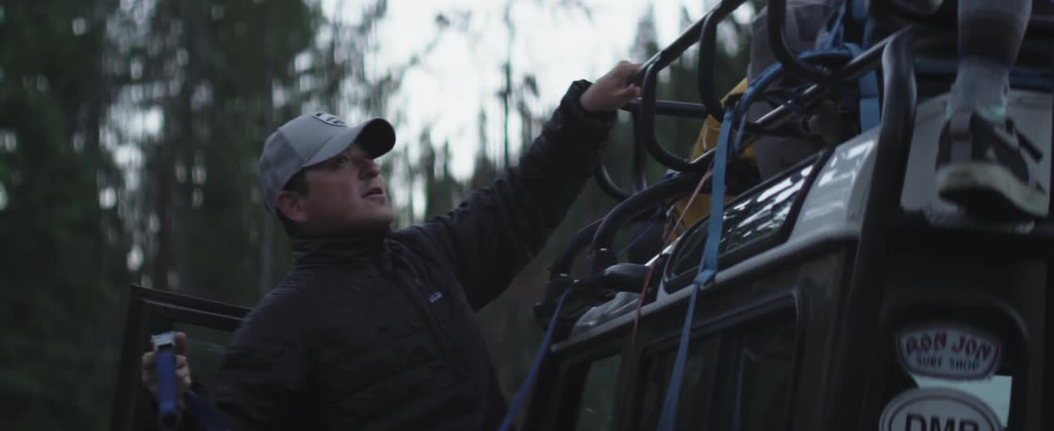 A man works with another to tie equipment to the top of a jeep-type vehicle