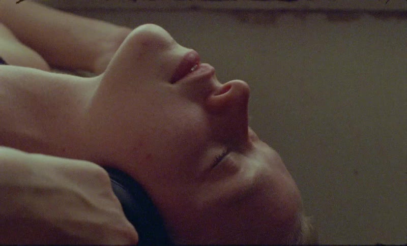 A woman wearing headphones lies with her head off the side of the bed swaying and listening to music