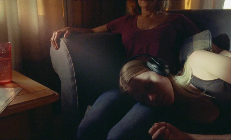 A girl wearing headphones lays down on a couch and puts her head on her moms lap