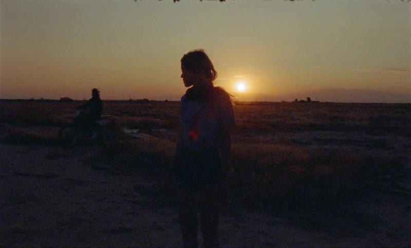 A woman stands on the field at dawn as a person rides their dirt bike past