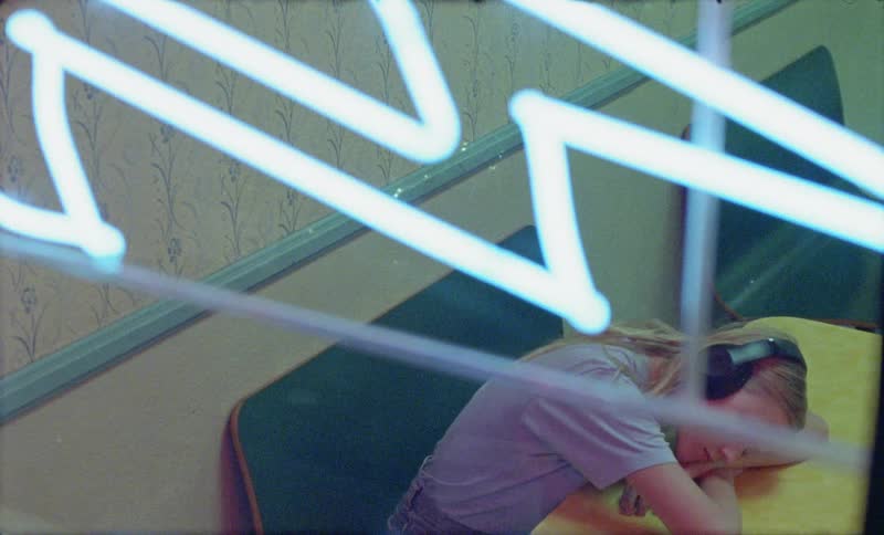 A neon sign glows on a diner window and inside a woman wearing headphones puts her head down on the table