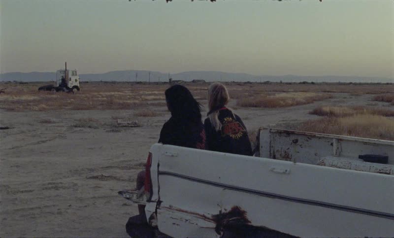 Two people sit on the back of a pickup truck looking towards the dry land area