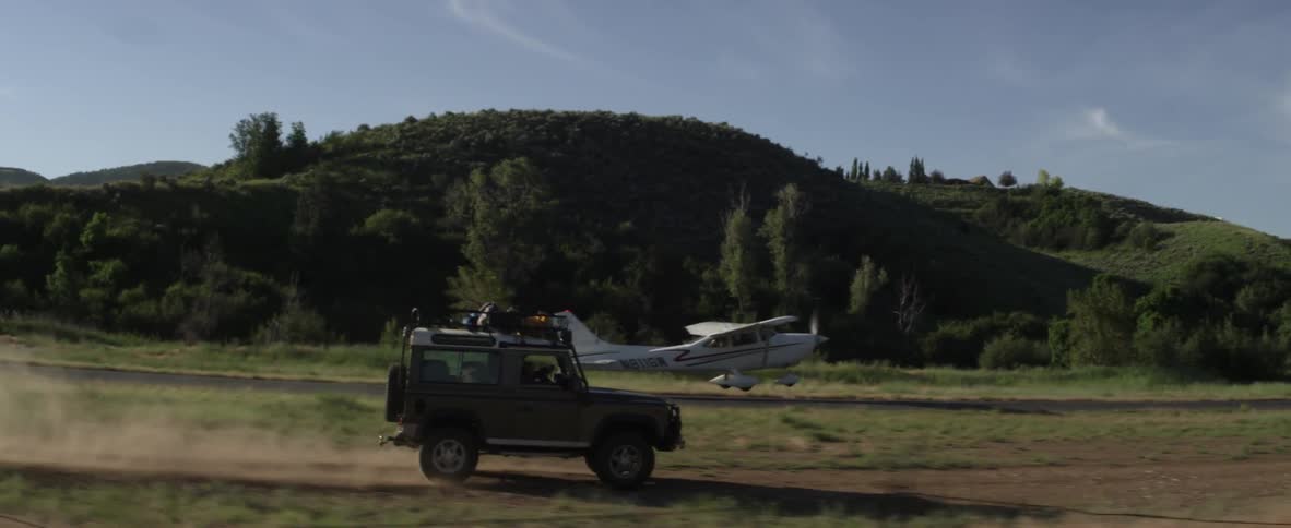 A jeep drives in a rural area as a small plane takes off next to it