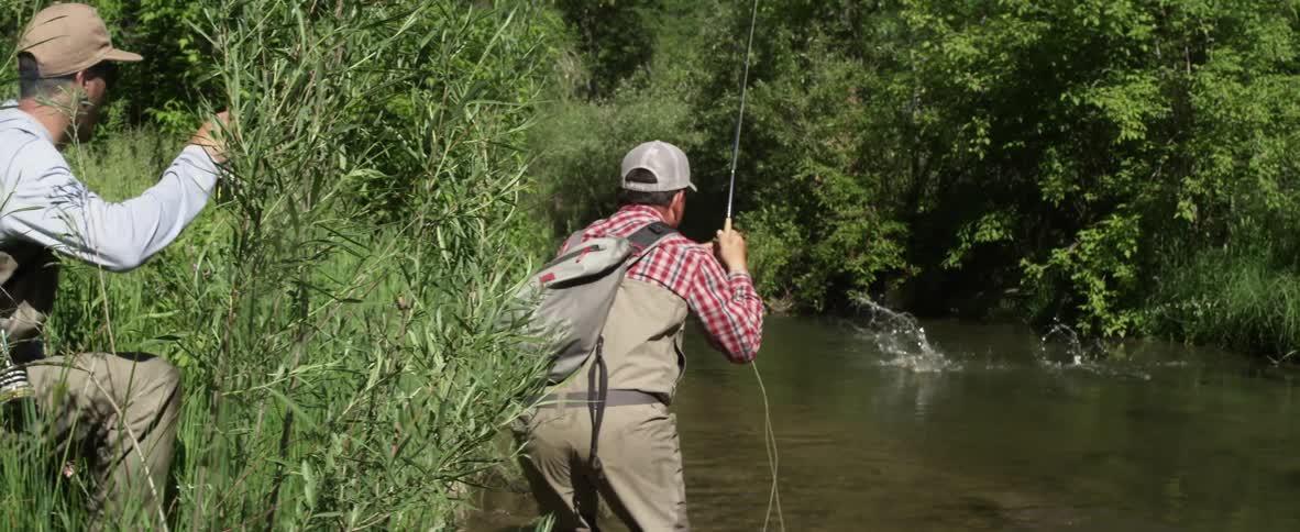 One fisherman standing at the edge of a body of water makes a catch with his rod as another fisherman looks on