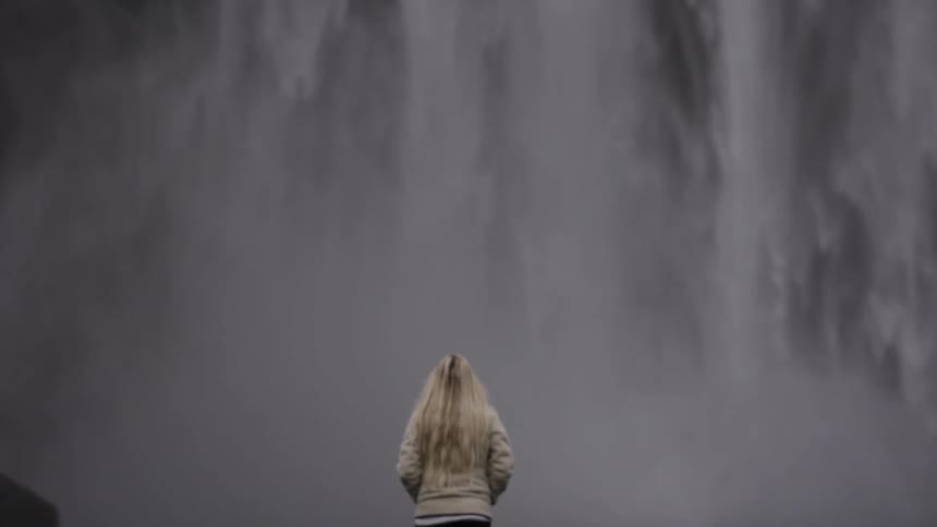 A blonde woman walks towards a violent waterfall