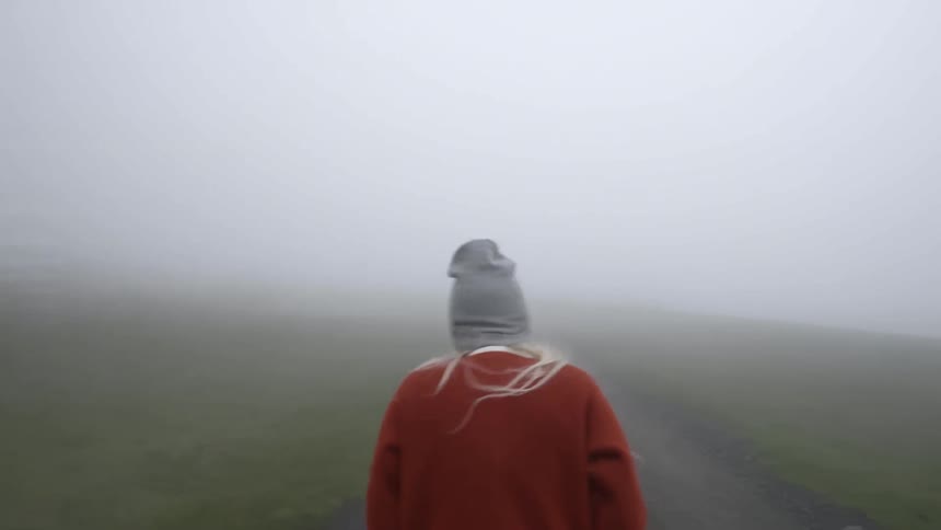 A blonde woman wearing a sweater and skullcap walks down a straight path in an open field on a very foggy morning
