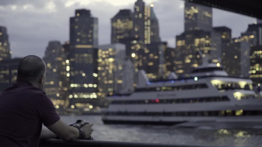 A man leaning over a railing as a river cruise ship passes between him and brightly-lit city skyline during dusk