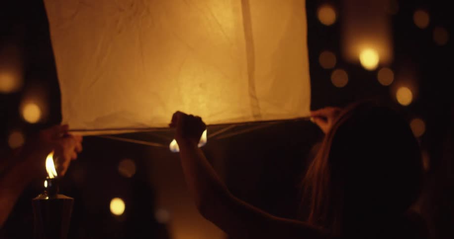 A girl and a man holding the edge of a chinese lantern as other lantern lights are seen floating in the background