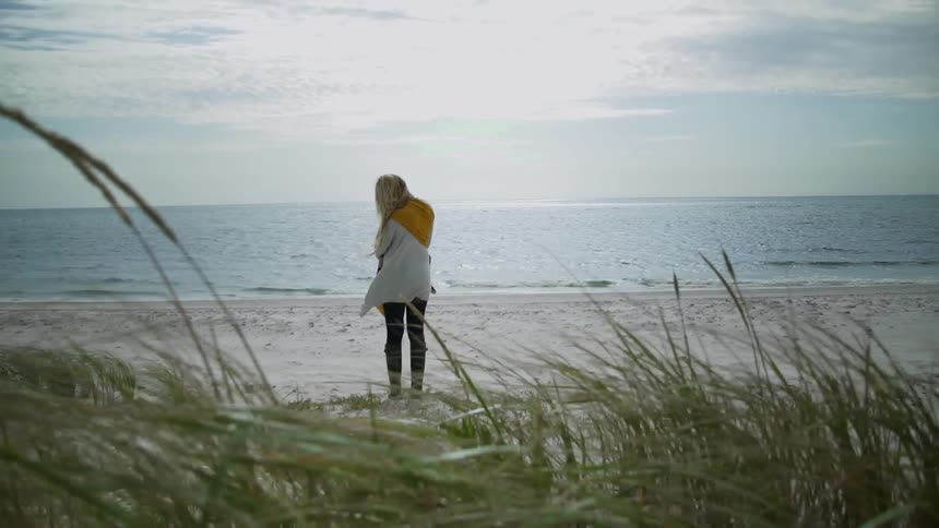 A woman with blond hair stands on the beach holding a baby