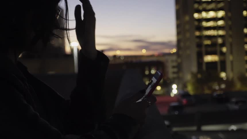 Woman stands on balcony looking at smartphone with city in the background