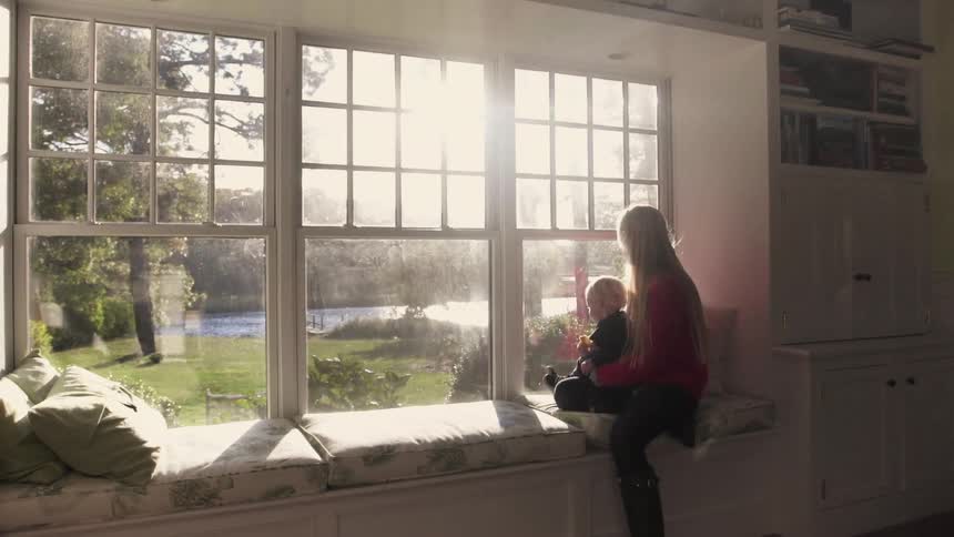 A woman carrying a toddler  towards a window seat and sitting down with him on her lap as they look outside o a sunny day