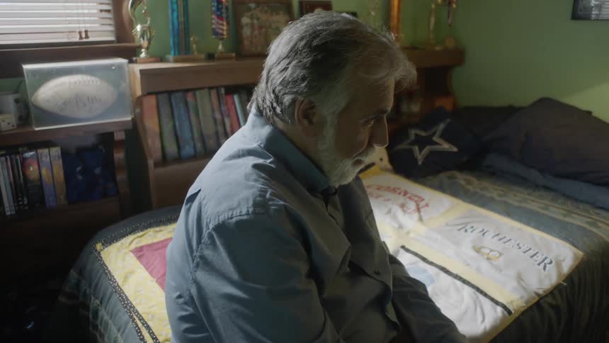 An old white man sits on a bed in a bedroom and cover his face with his hands