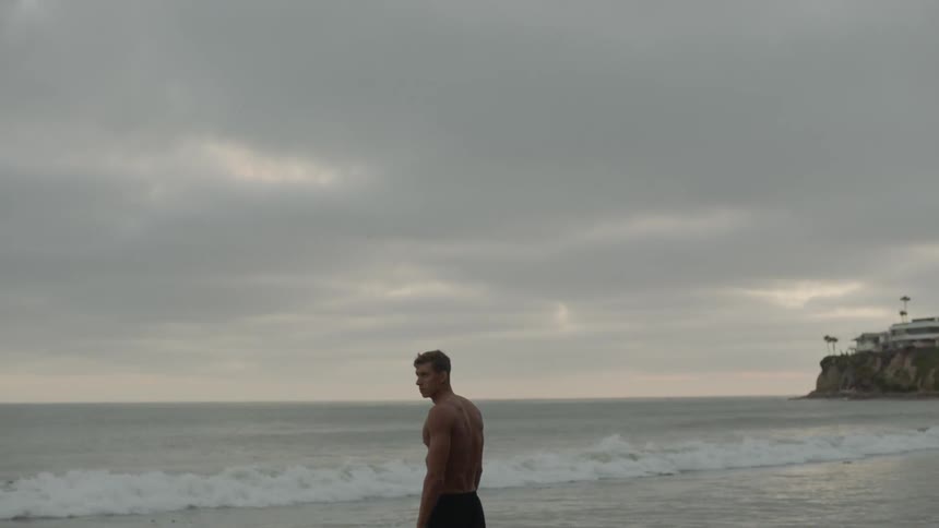 Shirtless muscular man stands on the beach next to the sea on a cloudy day