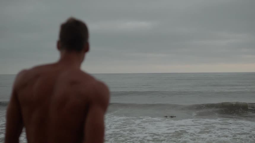 Shirtless muscular man walks into the sea on a cloudy day
