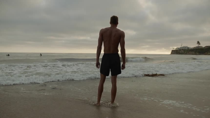 A shirtless man stands on the beach at dawn