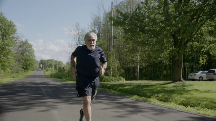 An older man with a white beard is jogging down a paved road in a rural area