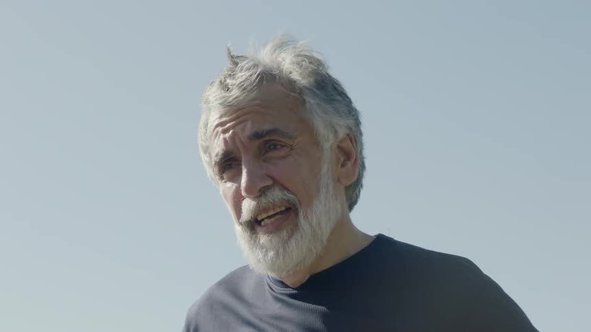 An elderly man stands outside in the wind looking off into the distance