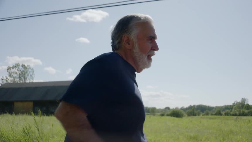 An elderly man with a white beard runs on a rural road