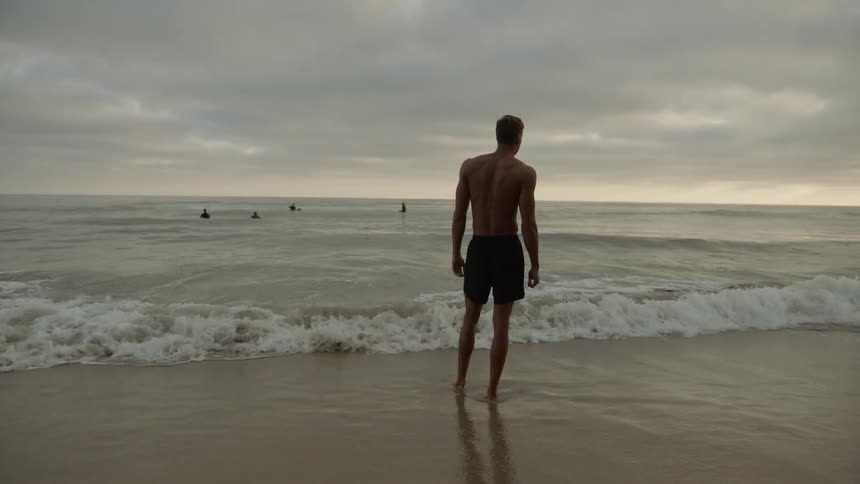 Muscular man stands on the sand beach next to the ocean