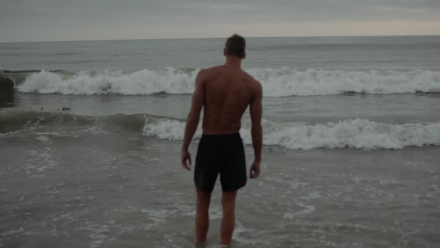 A white male in swimming trunks is standing ankle deep in water as the ocean tide rolls in