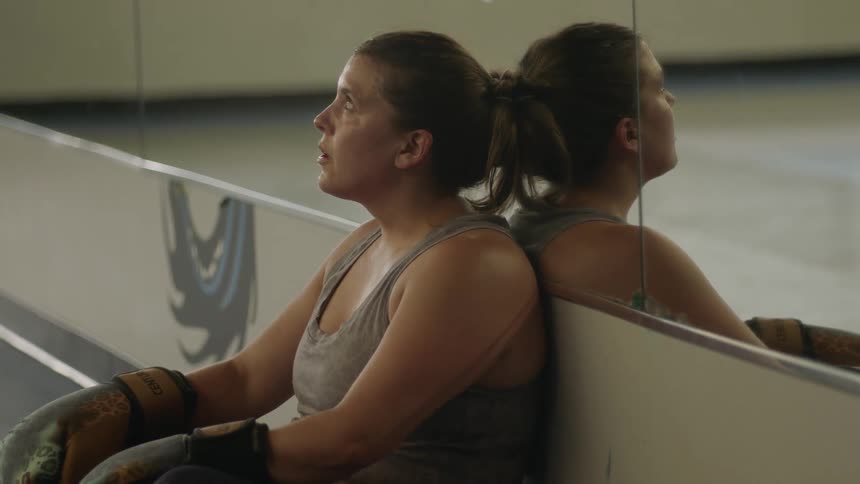 Sweaty female boxer sits on the ground and leans back against the wall and mirror in the gym