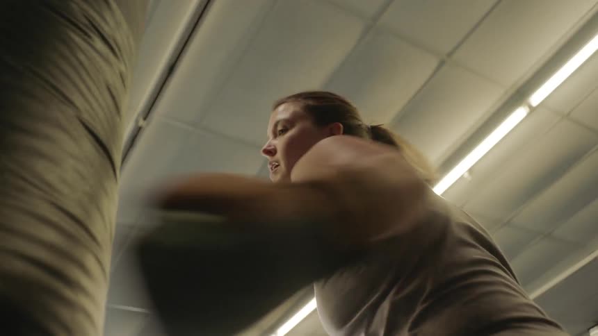 A white female is wearing boxing gloves to punch a punching bag in a large open space lights overhead