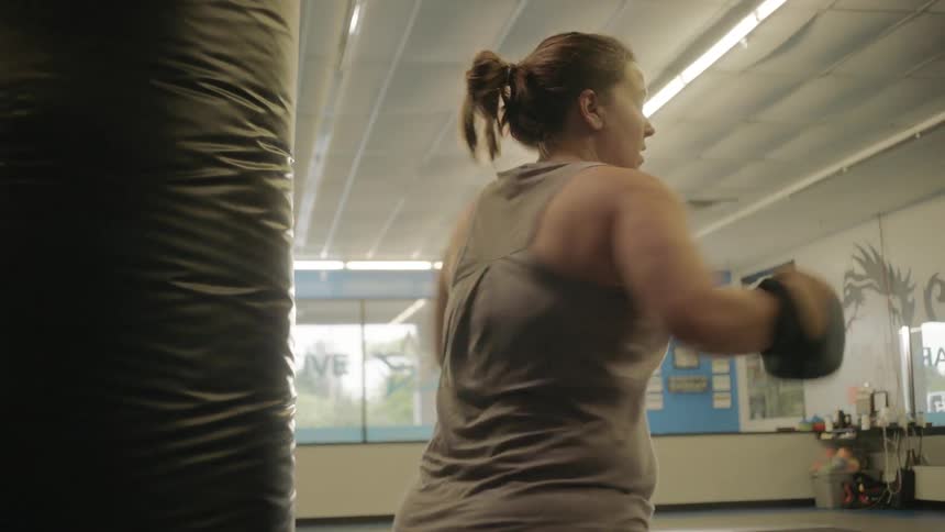 A white female is standing with her back to a punching bag and using boxing gloves to punch it behind her rotating back and forth between arms