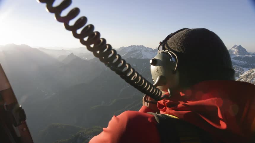 A man with a camera and a headset on a helicopter with mountains in the background