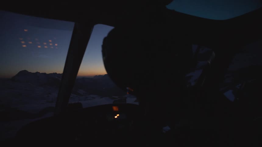 A pilot sits in the darkened cockpit of a helicopter as the sun has just dropped below the horizon behind some mountains