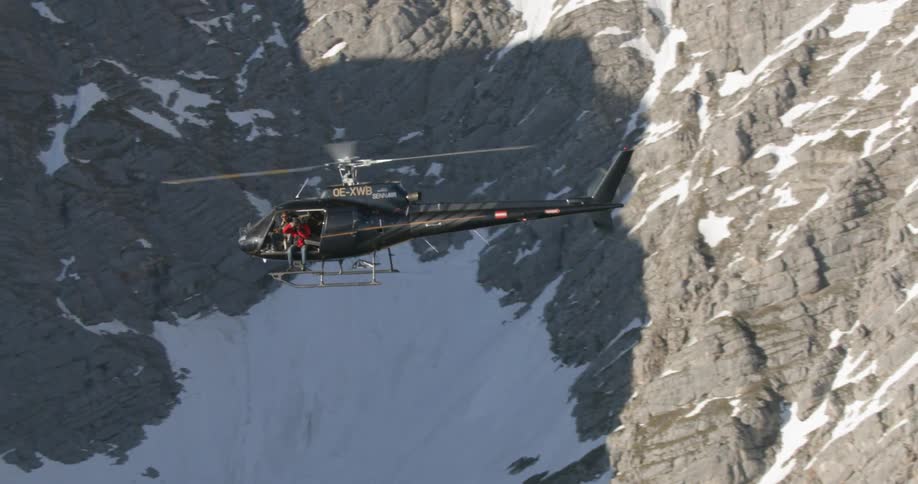 Cameraman leans out of helicopter as it flies over snow covered mountain slopes