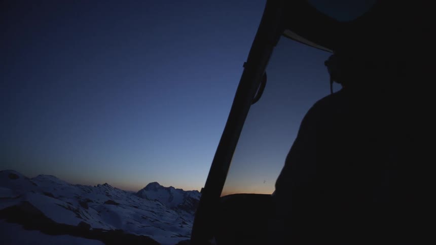 From within the cockpit of a helicopter the pilot looks out over the snow capped mountains in the distance as the sun has set below the horizon