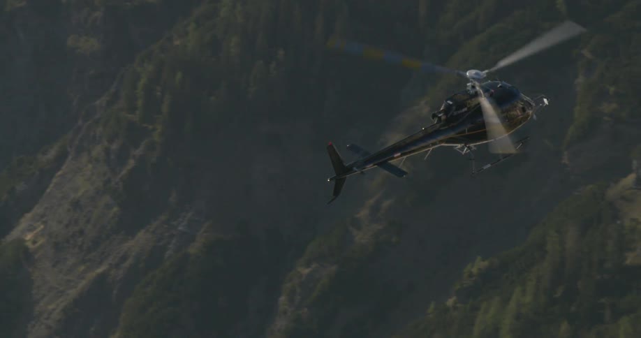 A dark colored helicopter flies high in the air over a forest covered mountain range and near a rocky cliff