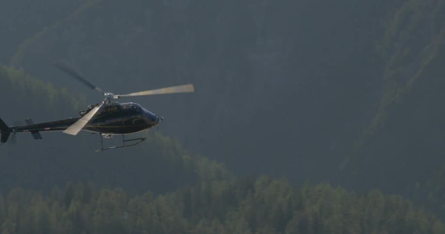 A helicopter flies over a forest and snow covered mountains