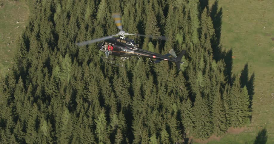 A helicopter with two men sitting on the edge flies over a forest