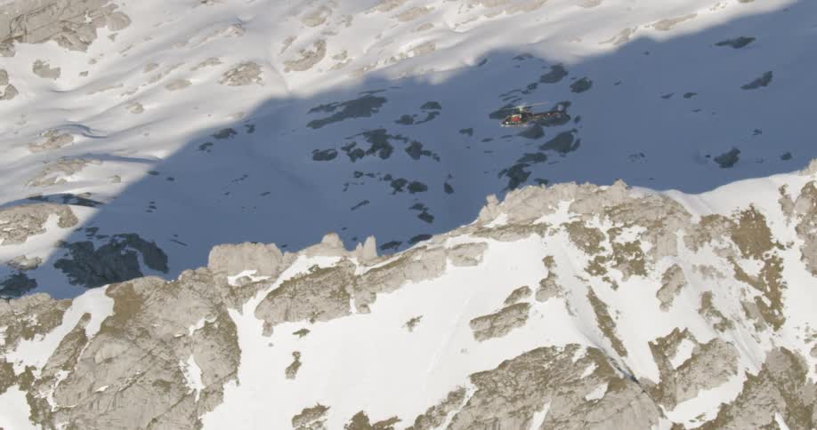 A helicopter flies over snow dusted rocky mountain ridges on a sunny day