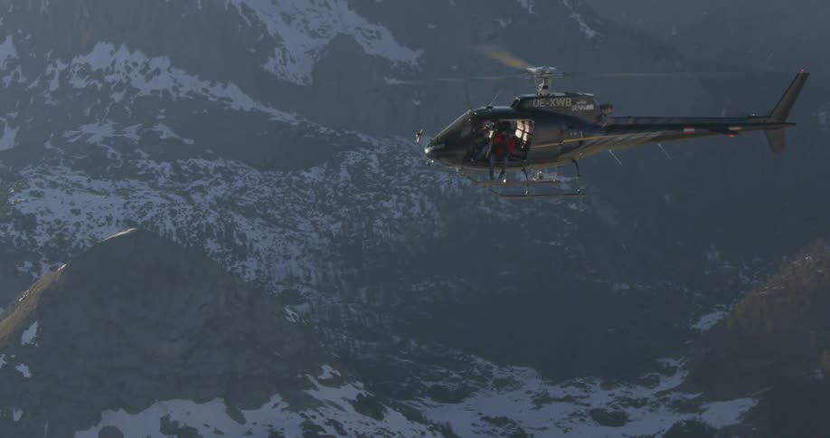 A person is sitting on the edge of a helicopter as it flies over a mountain range
