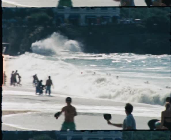 On a crowded beach people play in the surf and walk on the beach while two young males play a game of paddle ball