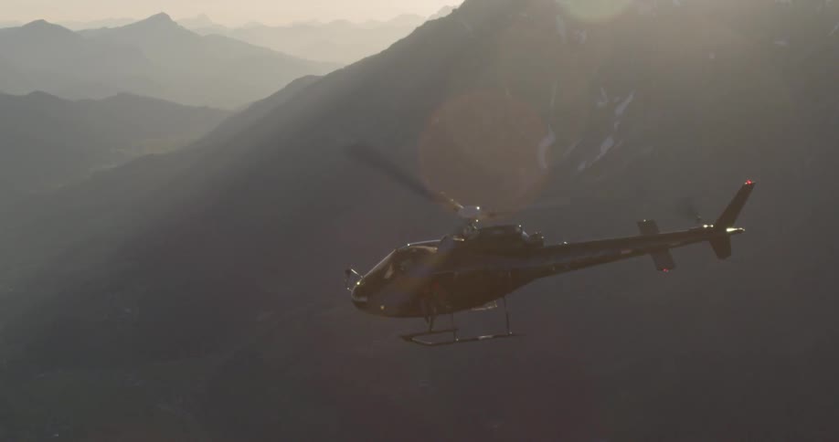 A helicopter with two people sitting on the edge flies through the mountains