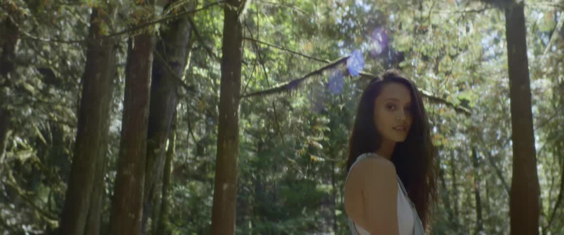 A woman standing in the trees near a boulder looks back over her shoulder