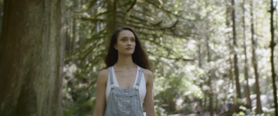 A thin female in jean short overalls is walking through a forest with the suns rays peeking through the tops of the trees