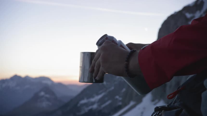 A man sits in the mountains and pours a drink out of his thermos