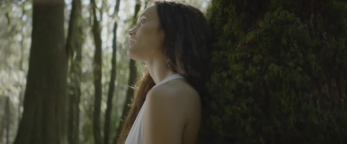 A brunette white woman is sitting and leaning against a tree trunk in a forest