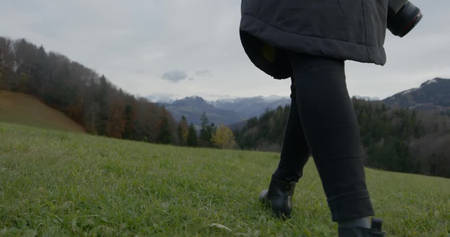 A woman wearing boots and a heavy coat walks along a grass hillside that is in the midst of a forest covered mountain range