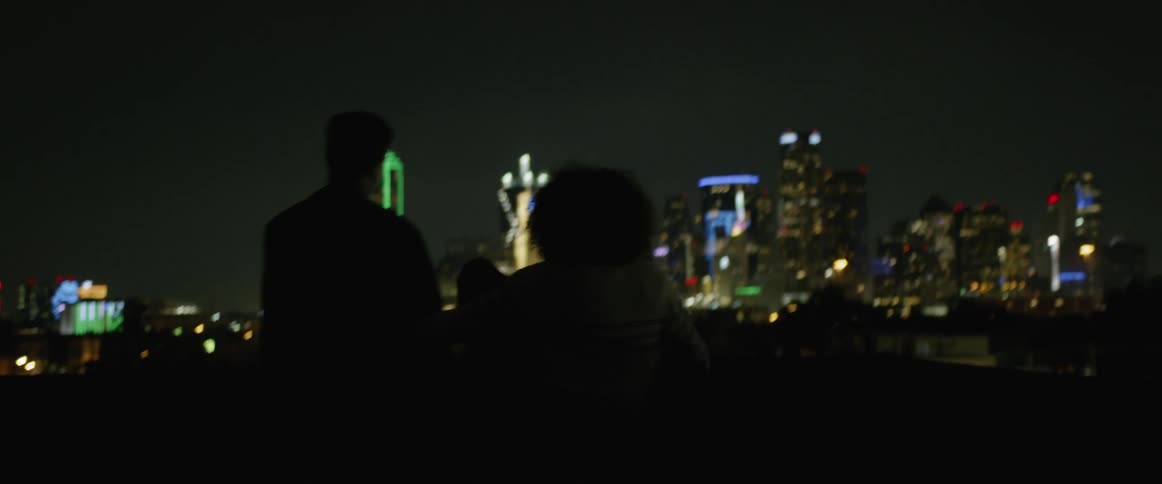 The silhouettes of a young man and woman can be seen against the brightly illuminated buildings of a downtown area of a major metropolis at night