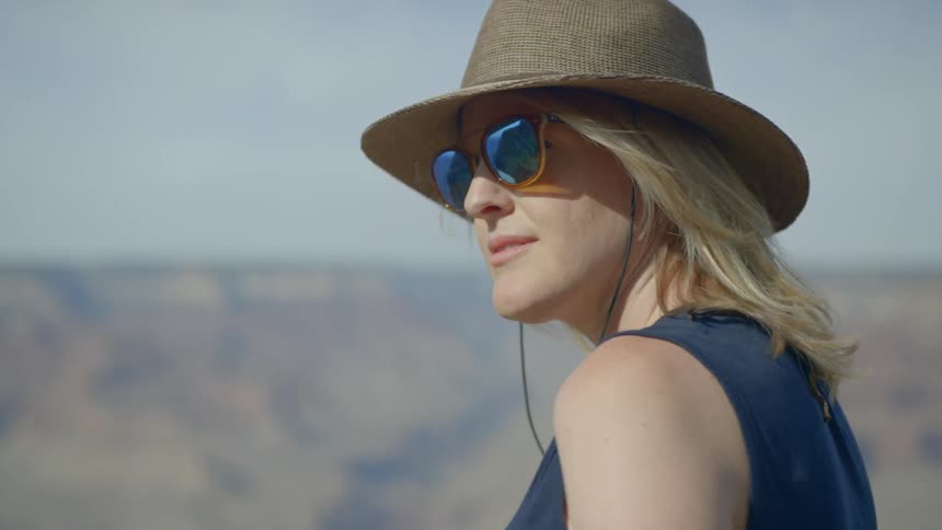 Blonde woman wearing wide hat and sunglasses looks across canyons during daylight