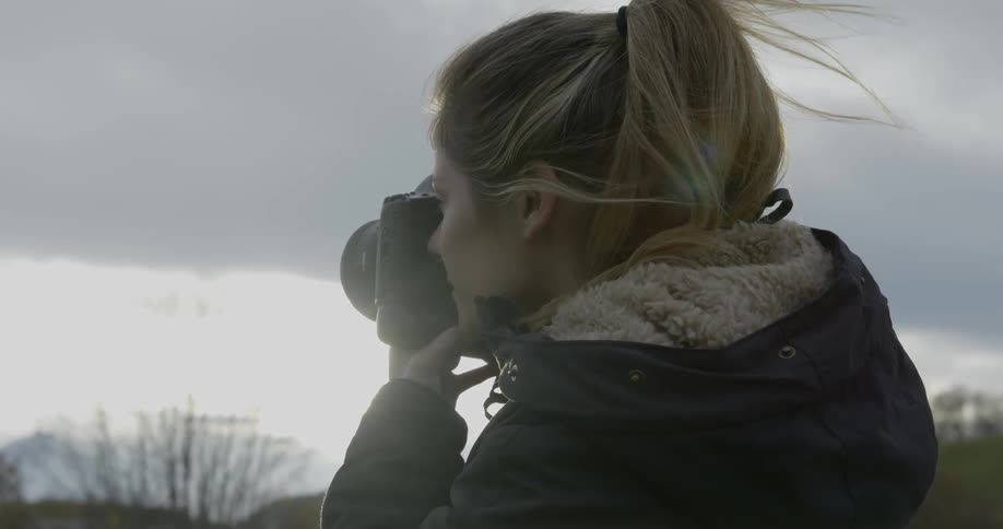 A blonde female is using a camera to take photos of something in the distance beneath a sky filled with dark clouds