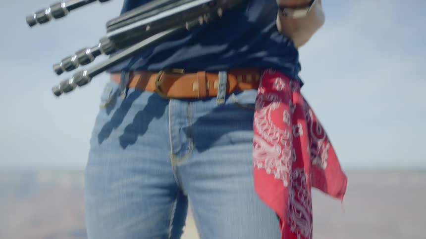 A woman who is standing outside in an arid environment reaches down and picks up a tripod and begins extending the legs